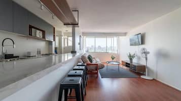 Uma cozinha moderna com uma ilha grande, uma sala de jantar com sofá e banquetas, e uma sala de estar com televisão e ventilador.