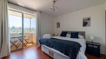 Quarto com cama, mesa de cabeceira, cadeira, mesa e vista da cidade através de uma porta de correr.