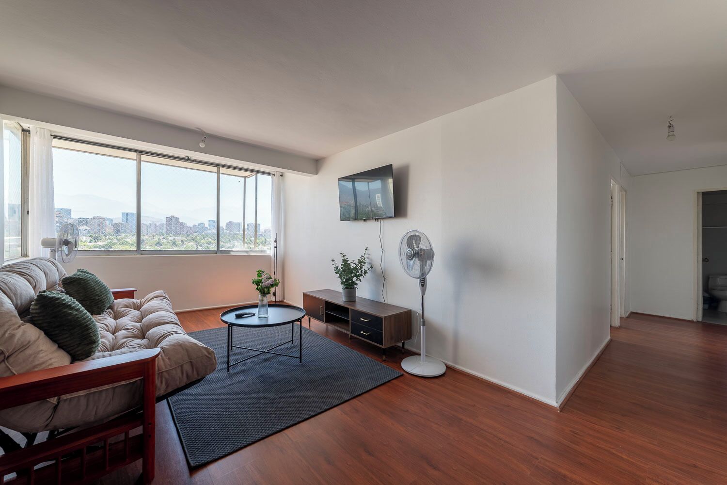 A modern living room with a sofa, a small table, a standing fan, and a TV mounted on the wall.