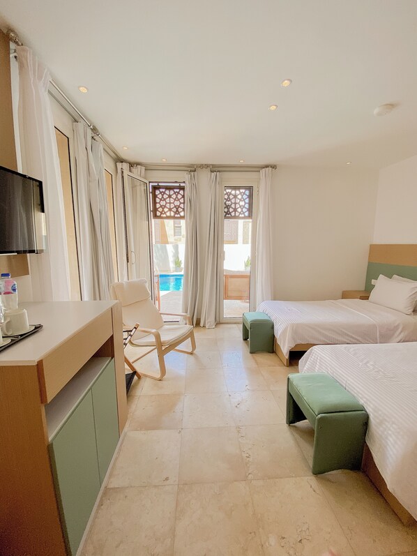 A hotel room with a desk, two beds, a chair, a TV, and a view of the pool.