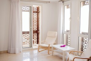 A room with a white chair, a small table with a purple flower, and a view through a wooden lattice door.