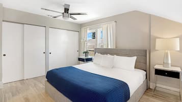 Een slaapkamer met een bed, nachtkastje, lamp, plafondventilator en raam met gordijnen.