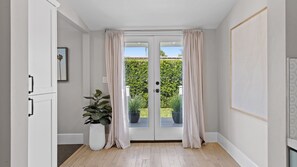 Un pasillo con un puerta doble que da a un jardín, una planta en maceta y un cuadro en la pared.
