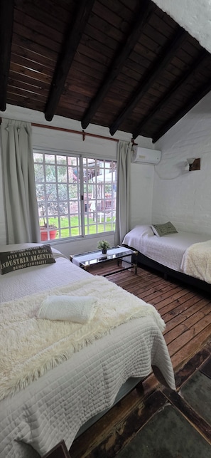 Ein Zimmer mit zwei Betten, einem fensterbelaubten Fenster, einem kleinen Tisch und einer weißen Wand.