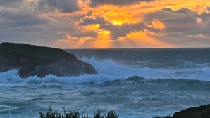 Un paisaje costero con olas rompiendo contra acantilados rocosos, una puesta de sol y un cielo nublado.