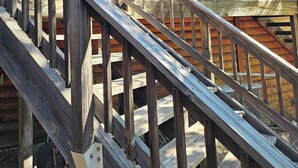Eine Holztreppe führt zu einem Balkon mit Geländer, einem Fenster mit Blick auf Bäume und einer Tür mit Fenster.