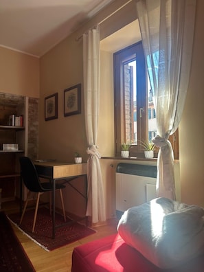 Ein Zimmer mit Schreibtisch, Stuhl und Bett. Vorhänge, ein Fenster mit Blick und eine Heizung sind vorhanden.