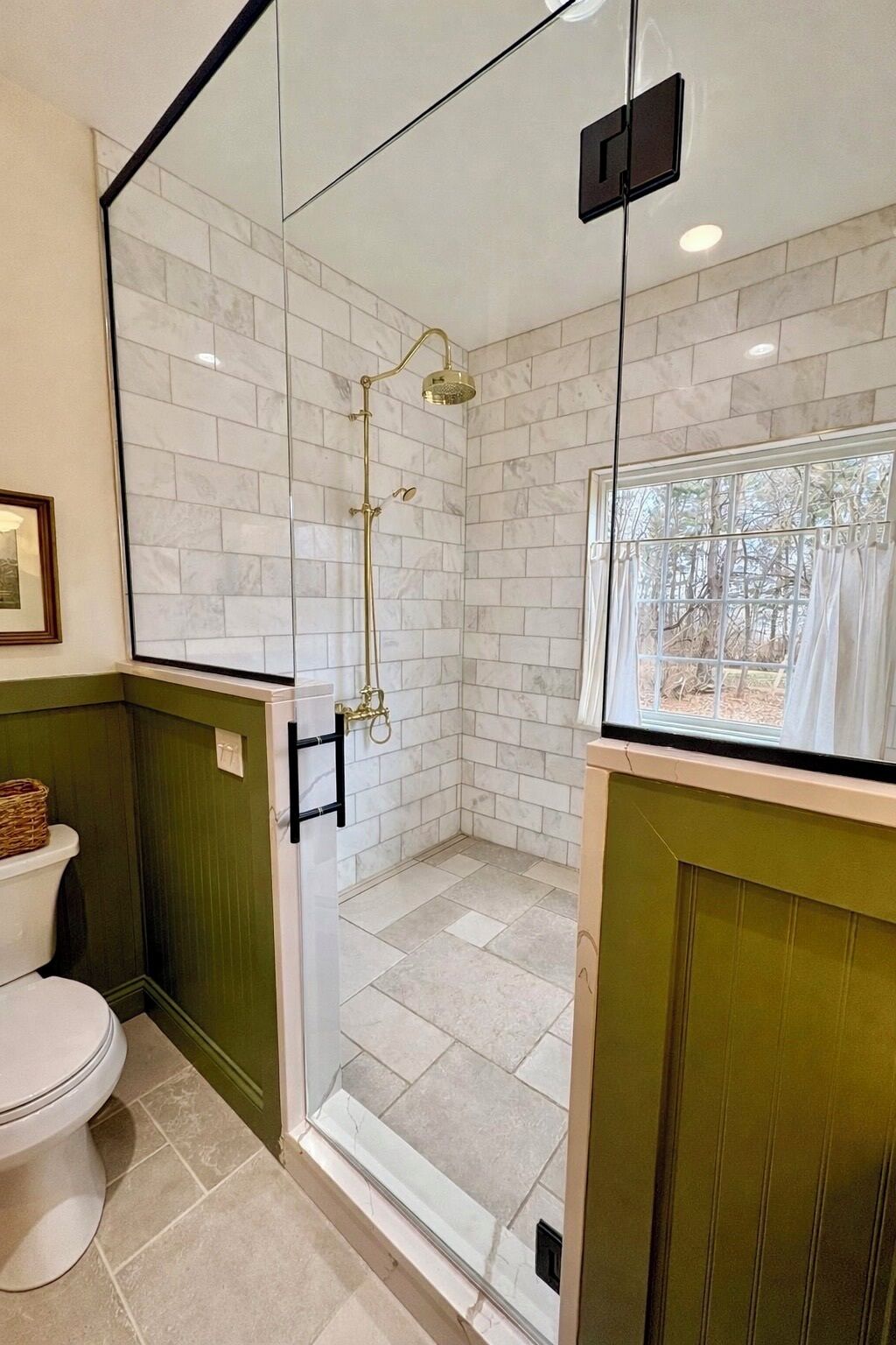 Casa de banho com chuveiro individualizado em vidro, sanita e janela com vista para árvores.