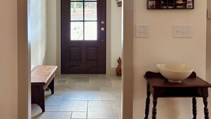 Um corredor com um banco de madeira, uma porta escura de madeira, piso ladrilhado, uma pequena mesa com uma tigela e uma prateleira com diversos objetos.