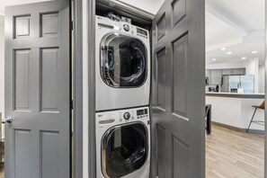 A laundry room with two stacked front-loading washers and dryers, a modern kitchen in the background, and a grey door. - Ski-in ski-out Beaver Creek W/pool and hot tub (Beaver Creek)