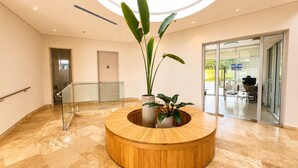 Uma área de recepção com balcão de madeira, plantas em vasos, porta de vidro que dá para uma área externa e guarda-corpo de vidro.