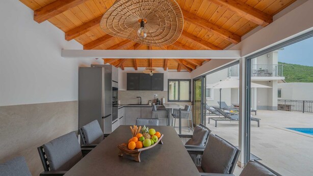 Eine moderne Küche mit Essbereich, einem großen Tisch voller Obst und Blick auf einen Pool sowie Sitzmöglichkeiten im Freien.