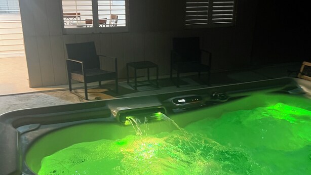 Ein Whirlpool mit grüner Beleuchtung, ein Fenster mit Jalousien und ein Stuhl draußen.