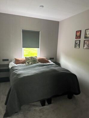 A bedroom with a bed, a window with blinds, a carpeted floor, and wall-mounted artwork. - Design House in Akureyri – Private Hot Tub, Sea & Mountain Views, 2 bathrooms (Akureyri)