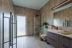 A modern bathroom with a glass shower enclosure, a toilet, a sink with a mirror, and a door leading to another room. - Design House in Akureyri – Private Hot Tub, Sea & Mountain Views, 2 bathrooms (Akureyri)