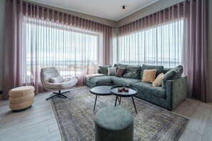 A modern living room with a sectional sofa, a round coffee table, a round ottoman, a chair, and a large window with curtains. - Design House in Akureyri – Private Hot Tub, Sea & Mountain Views, 2 bathrooms (Akureyri)