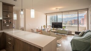 Un appartement moderne comprenant une cuisine, un coin repas et un salon, et doté d’une grande fenêtre offrant une vue sur la ville.