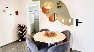 Uma sala de jantar moderna com uma mesa redonda, cadeiras estofadas cinzentas, uma luminária pendente e uma cozinha visível ao fundo.
