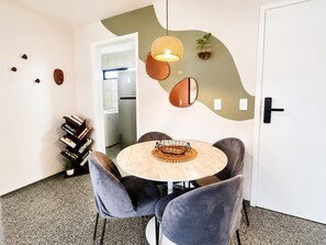 A modern dining area with a round table, grey upholstered chairs, a hanging light fixture, and a kitchen visible in the background. - Hosts BR - Ap. com Rooftop Vista Mar (Fortaleza)