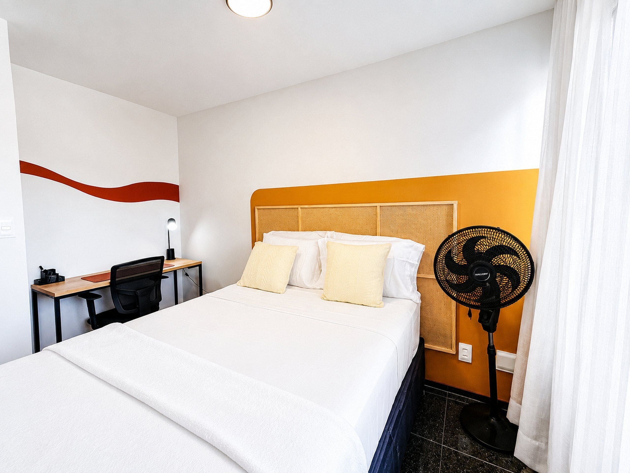 Une chambre d’hôtel comprenant un lit, un bureau, une chaise, un ventilateur sur pied et une salle de bain.