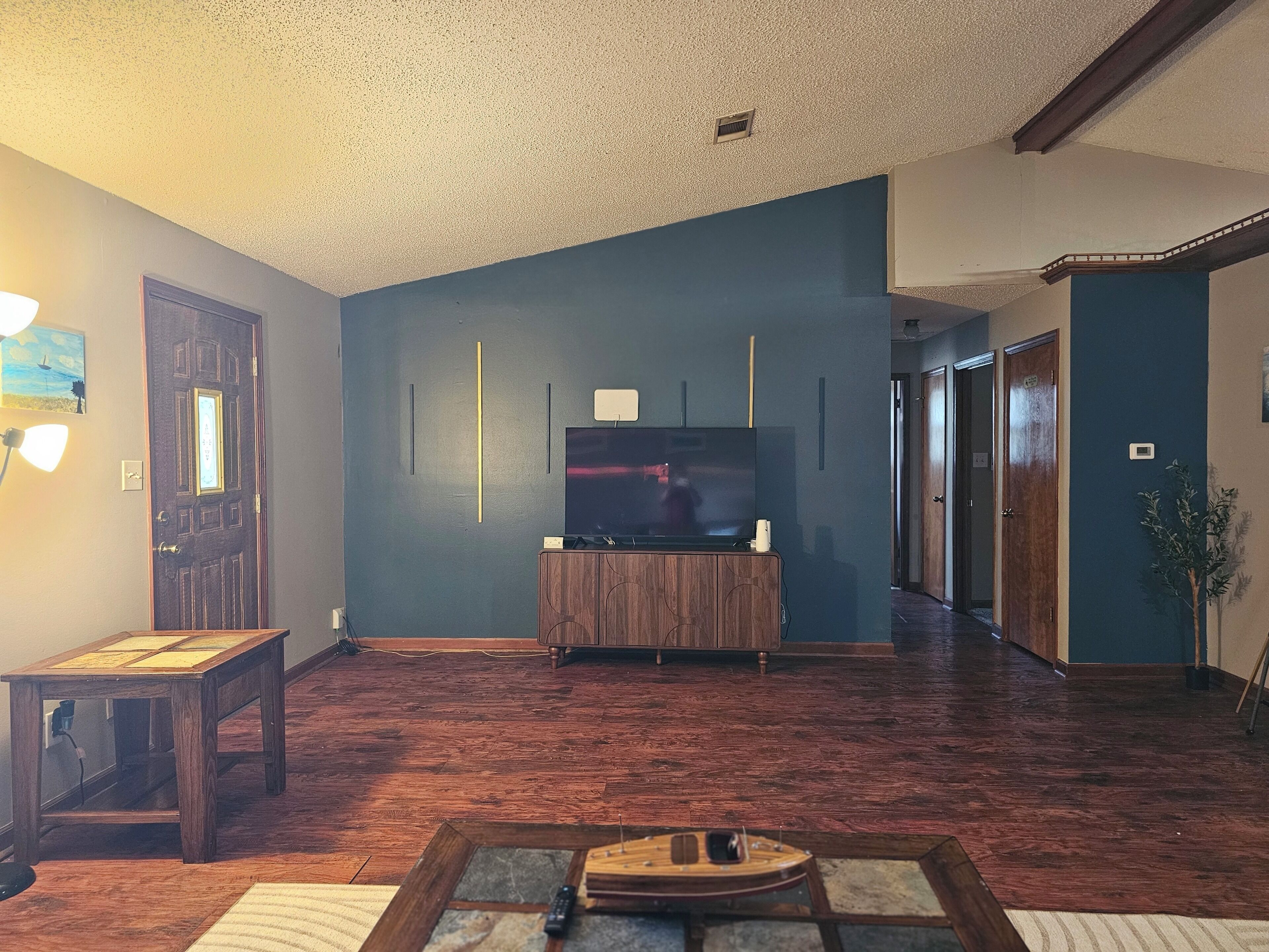 Un salon avec un plancher en bois, un téléviseur posé sur un meuble en bois et une porte dotée d’un luminaire.