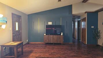 Un salon avec un plancher en bois, un téléviseur posé sur un meuble en bois et une porte dotée d’un luminaire.