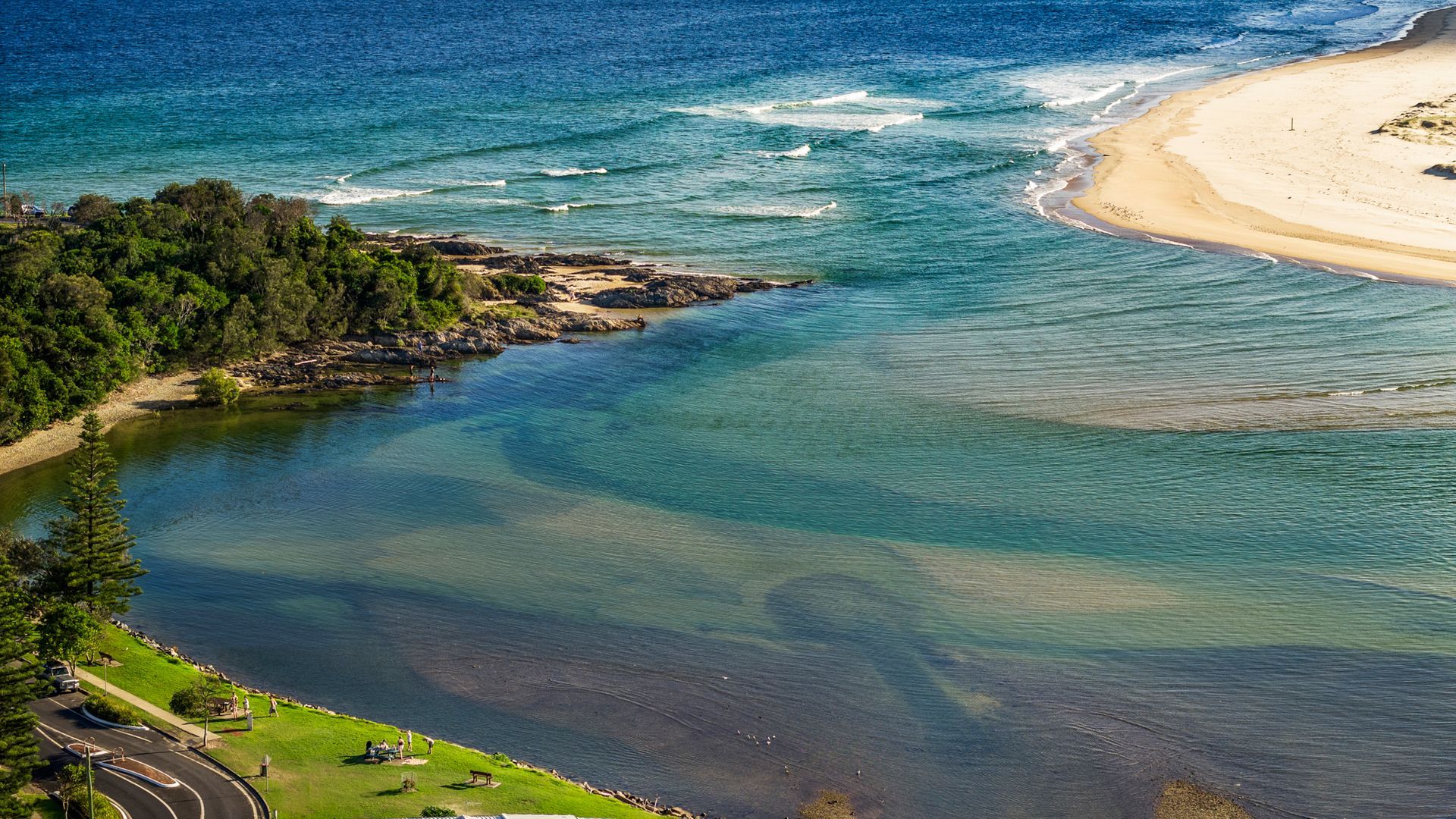 A coastal landscape with a beach, clear water, and a road leading to a grassy area.