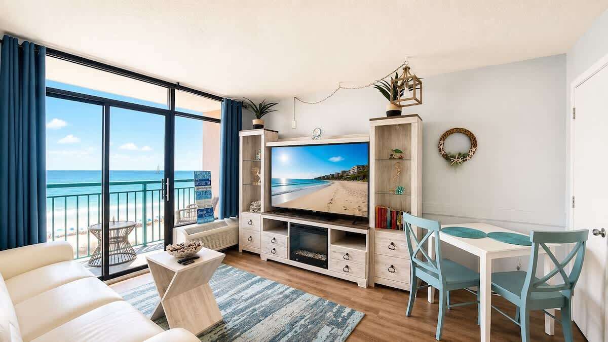 Ein Wohnzimmer mit Fernseher, Esstisch, Stühlen, einer Couch, einem Balkon mit Meerblick und einem kleinen Tisch mit einer Schüssel.