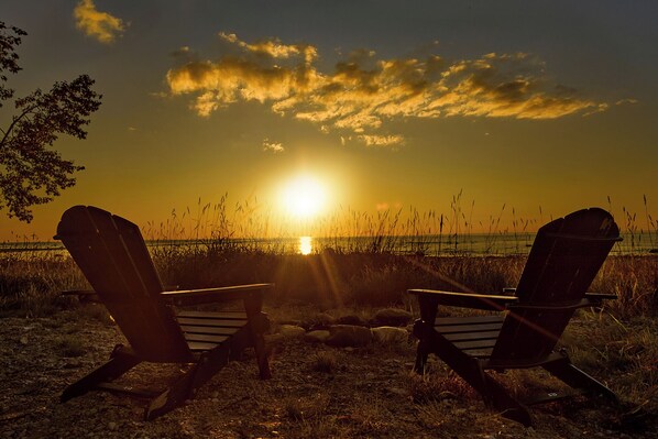 Zwei Adirondack-Stühle, die auf einen Sonnenuntergang über dem Wasser zeigen.