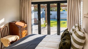 Ein Schlafzimmer mit einem Bett, einer Holzkommode, einem Sessel und Blick durch eine Glastür in einen Außenbereich.