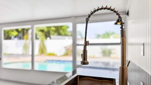 Ett kök med en modern blandare, en rektangulär diskho och utsikt över en swimmingpool genom stora fönster.