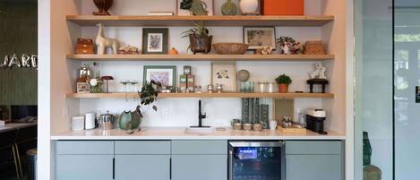 A kitchen with a refrigerator, shelves with decorative items, and a dining area visible in the background.