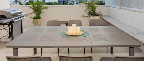A rooftop terrace with a table, chairs, a grill, and potted plants.