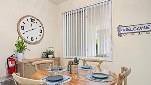 Un comedor con una mesa de madera redonda, cuatro sillas, un reloj de pared, un cartel de bienvenida y una ventana con persianas.