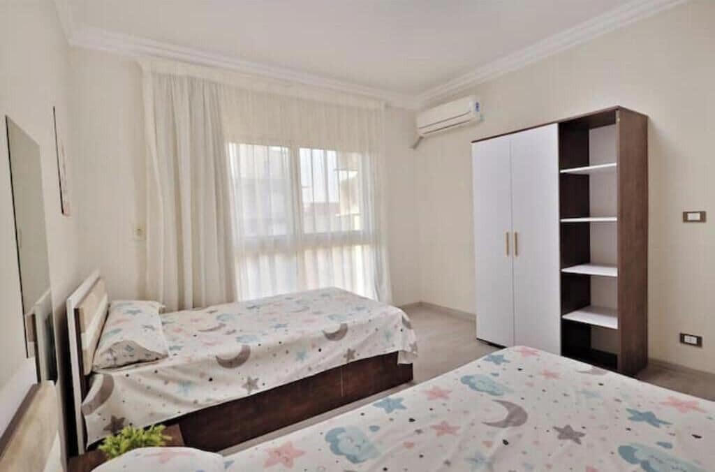 Quarto com duas camas, um guarda-roupa e uma janela com cortinas.