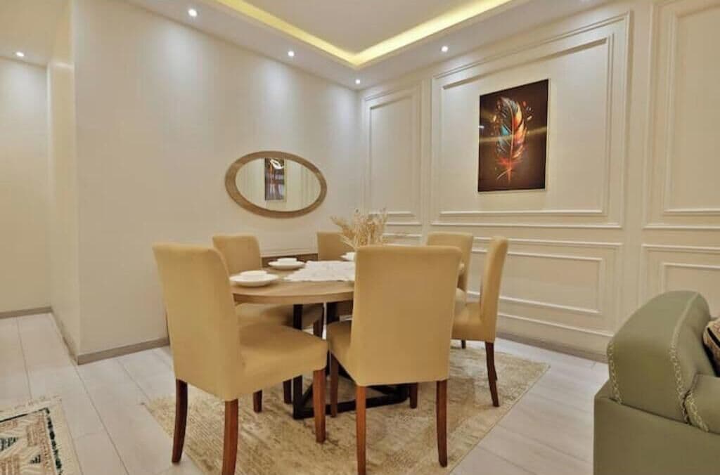 Uma sala de jantar com uma mesa redonda, seis cadeiras, um espelho e uma obra de arte emoldurada na parede.