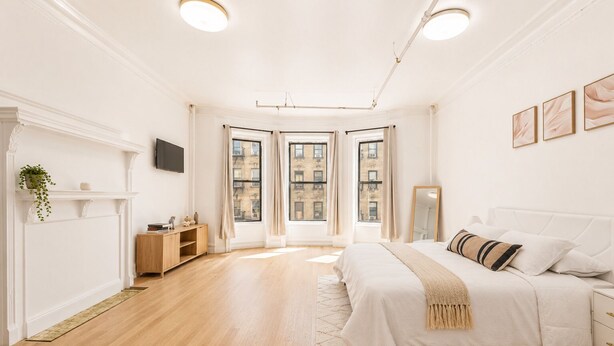 A bedroom with a bed, a nightstand, a dresser, a mirror, and a fireplace. - Bright Uptown Manhattan Studio Near Subway and Dining (New York)