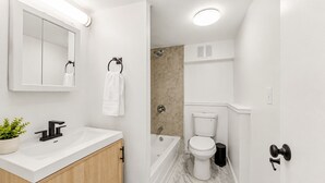 A modern bathroom with a white sink, mirror, and toilet, a shower area, and a potted plant on the counter. - Bright Uptown Manhattan Studio Near Subway and Dining (New York)