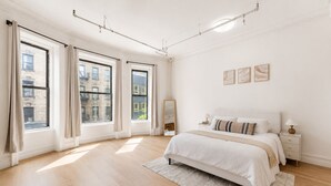 A bedroom with a large bed, a nightstand, a mirror, and a view of buildings through large windows. - Bright Uptown Manhattan Studio Near Subway and Dining (New York)