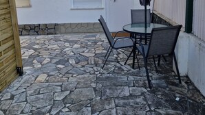 Um pátio com piso de pedra, uma mesa com cadeiras, uma porta e uma janela com persianas.