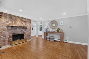 A room with a brick fireplace, wooden flooring, a white door, and a console table with decorative items. - Cape Cod Escape | Near Beaches | Play Area (Osterville)