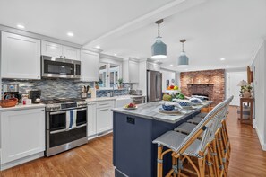 A modern kitchen with white cabinets, a central island, and a brick fireplace. - Cape Cod Escape | Near Beaches | Play Area (Osterville)
