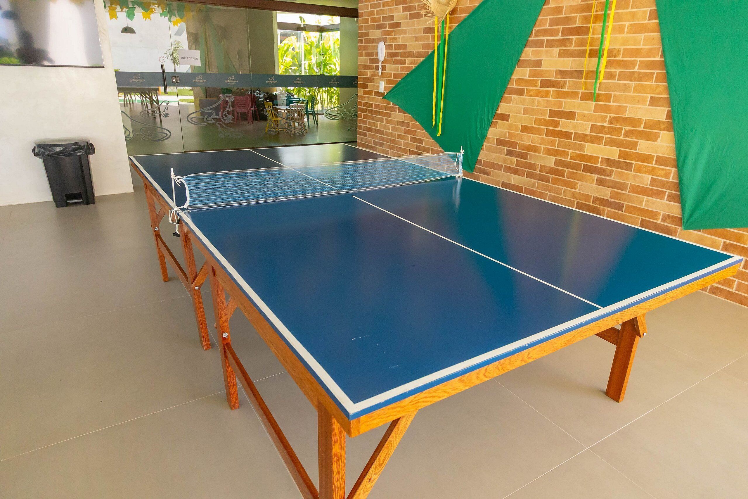 Ein Tischtennis-Tisch mit blauer Spielfläche und Holzbeinen, platziert in einem Raum mit Backsteinwand und Glastür zum Außenbereich.