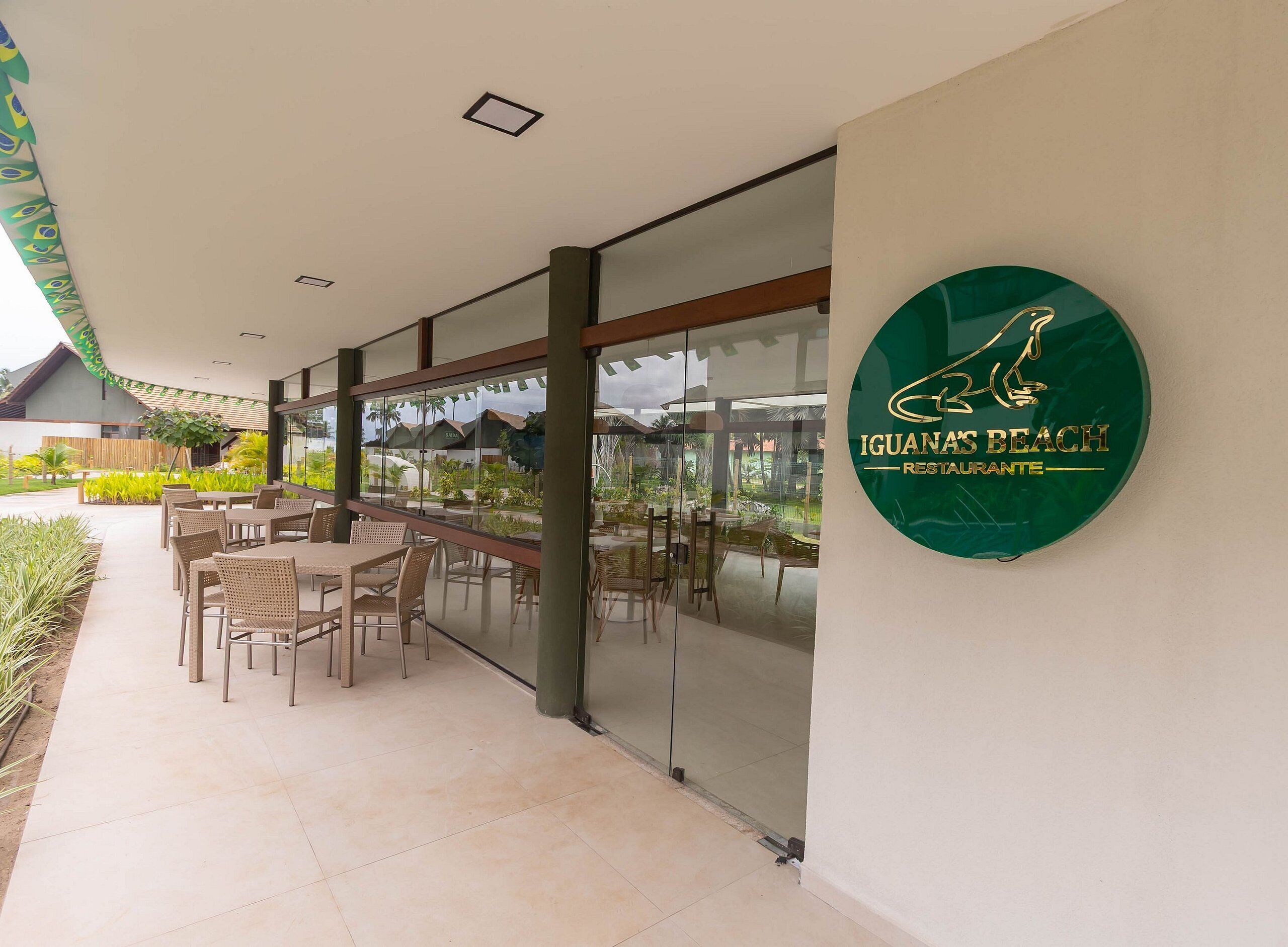 Un área de comedor al aire libre con mesas y sillas, un muro de vidrio y un letrero para Iguanas Beach Restaurante.