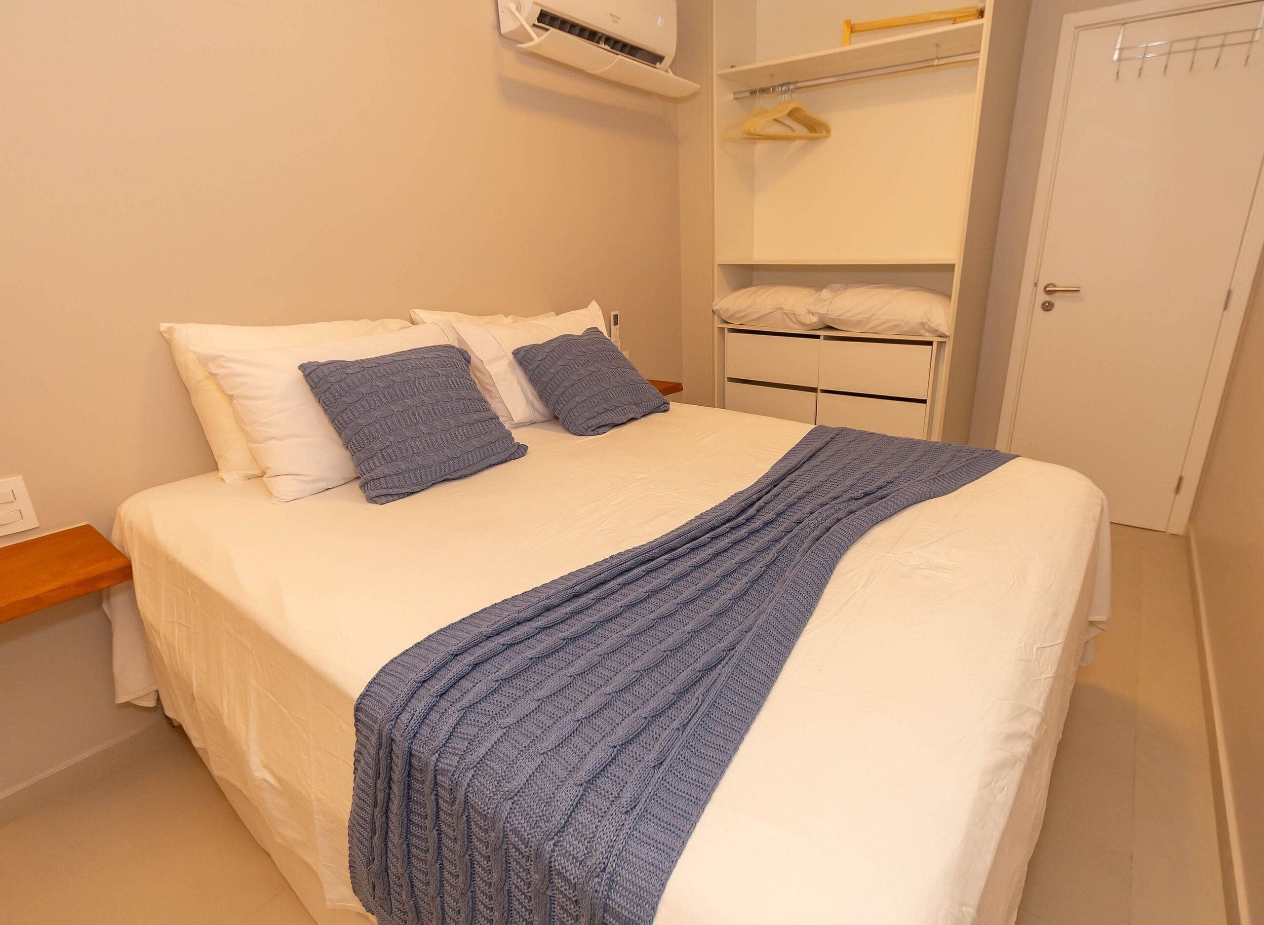 Una cama bien hecha con una manta y almohadas azules, una mesita de noche de madera, una pared blanca y una puerta blanca.