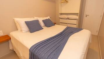 Una cama bien hecha con una manta y almohadas azules, una mesita de noche de madera, una pared blanca y una puerta blanca.