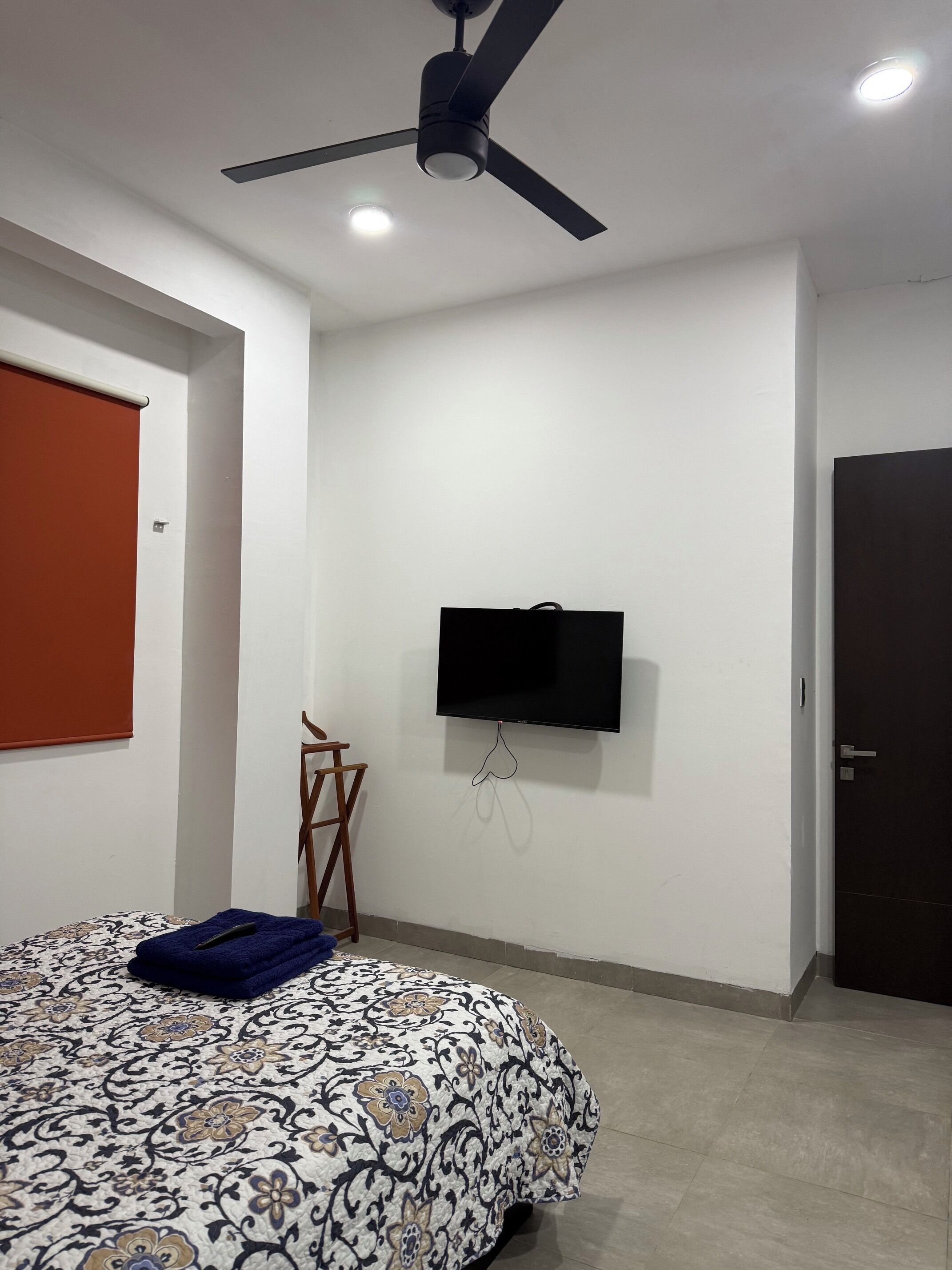 Un dormitorio con cama, ventilador de techo, televisor montado en la pared y una puerta.
