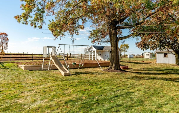 A playground with swings and a slide, a small white building, and a tree in a grassy field. - 3 Bedroom House - Family Friendly - Sleeps 6 (Ephrata)