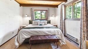 Un dormitorio con techo de madera, un ventanal con cortinas, cama con almohadas, una mesita de noche y un banco.