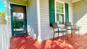 A covered porch with a red wooden floor, two chairs, and a window with green shutters.